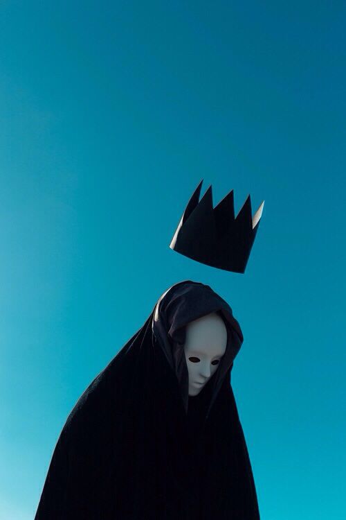 a person wearing a black cloak with a crown on their head and a blue sky in the background