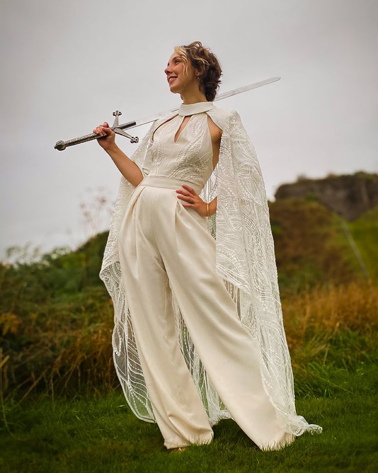 a woman dressed in white holding two swords and posing for the camera with grass behind her