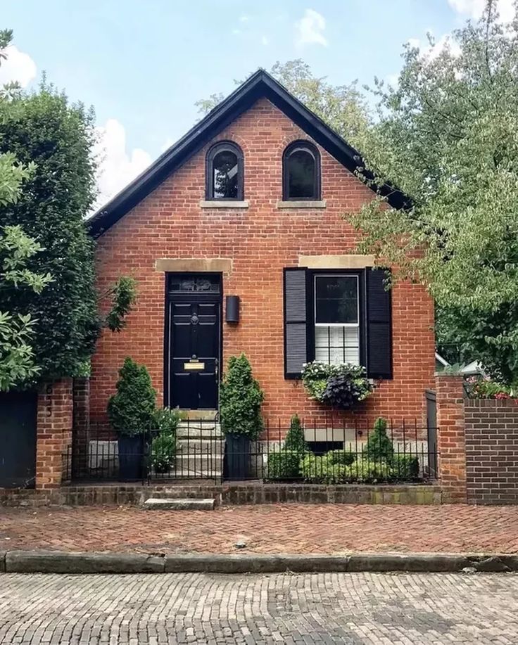 a brick house with black shutters on the front door and windows, surrounded by trees