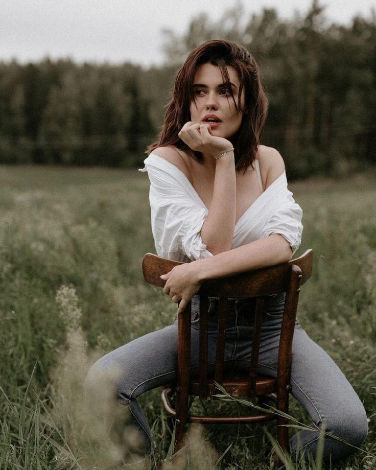 a woman sitting on top of a wooden chair in a field with her hand under her chin