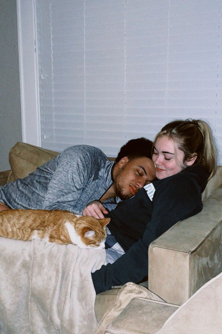 a man and woman laying on a couch cuddling with an orange tabby cat