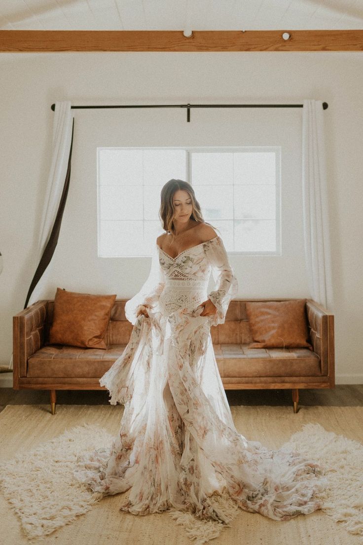 a woman standing in front of a couch wearing a dress with long sleeves and flowers on it