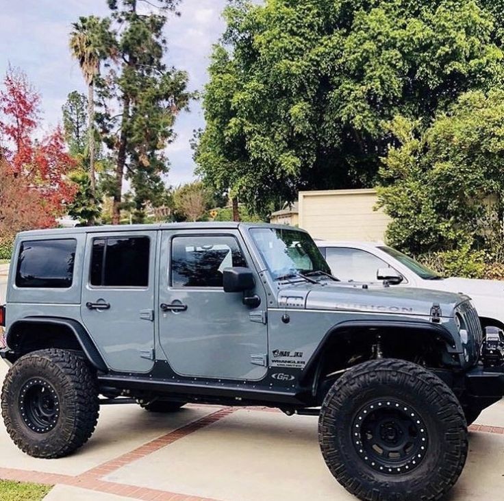 a gray jeep parked in front of a white truck on a driveway next to trees