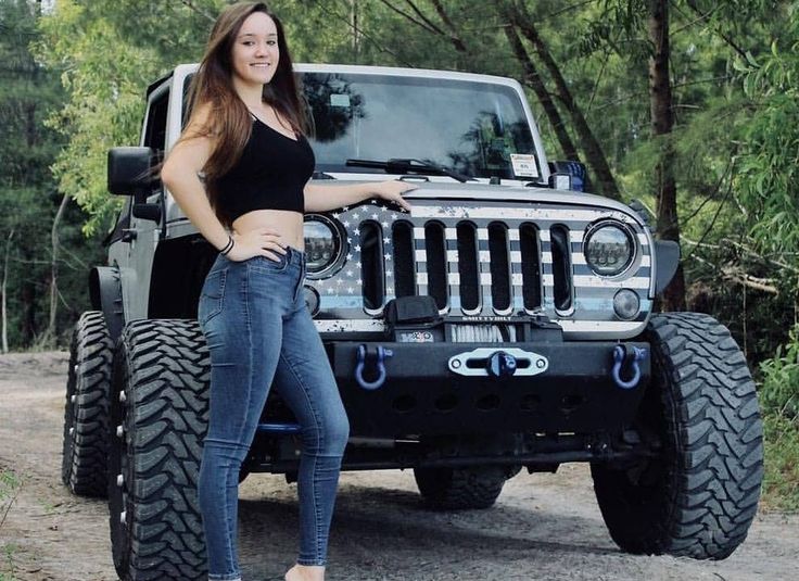 a beautiful young woman standing next to a black jeep parked in the woods with her hands on her hips