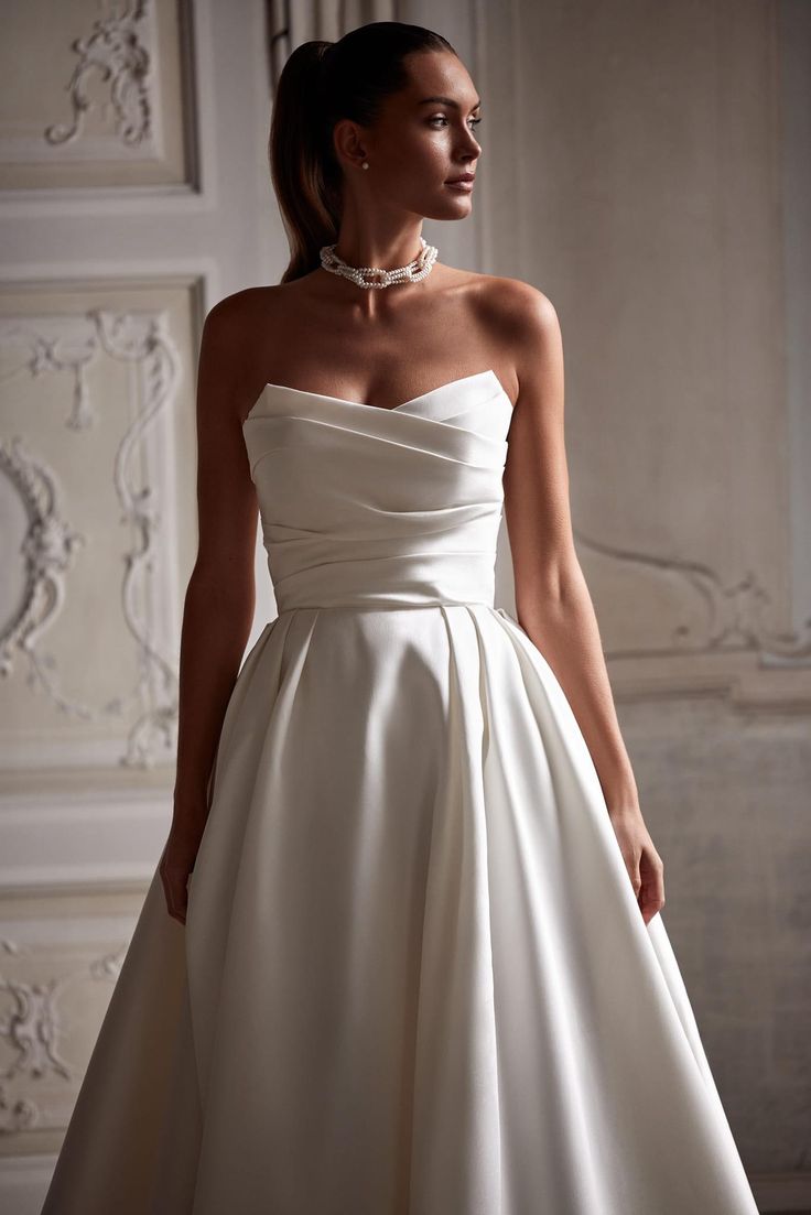 a woman in a white wedding dress with pearls on her neck and shoulders, standing next to a wall