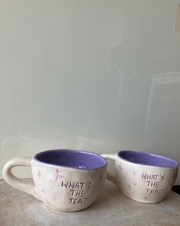 two white cups with purple rims that say what's the tea?