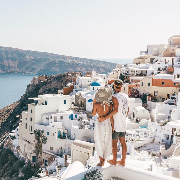 two people standing on the edge of a cliff with white buildings and blue water in the background