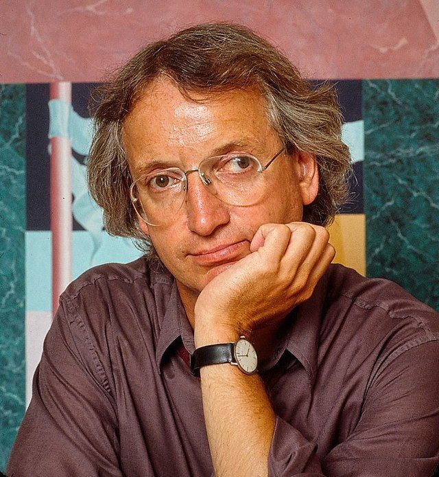 a man with glasses and a watch on his wrist sitting in front of a wall