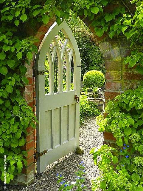 an open white door surrounded by greenery in a stone walled garden with gravel path