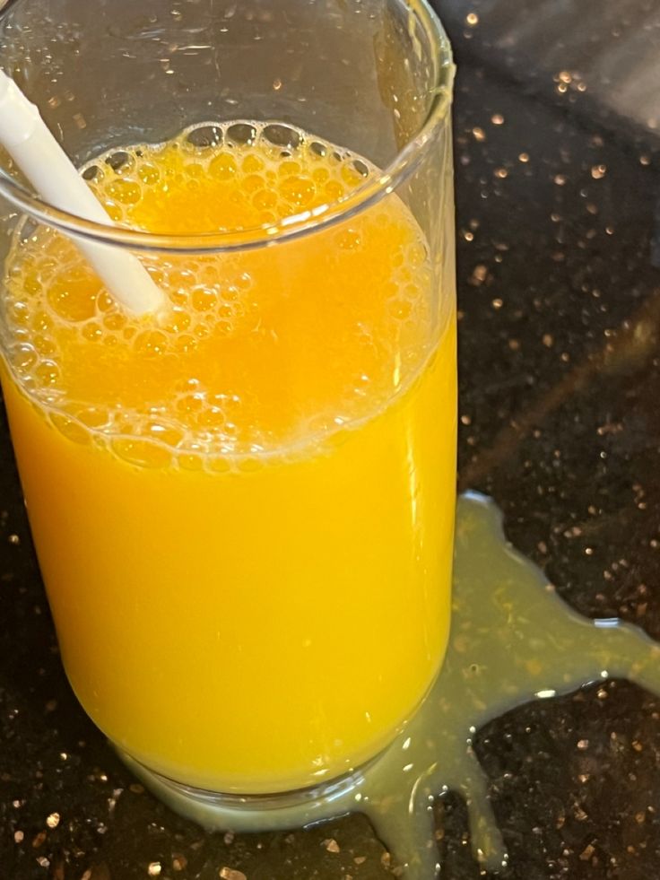 a glass filled with orange juice on top of a black counter next to a white spoon