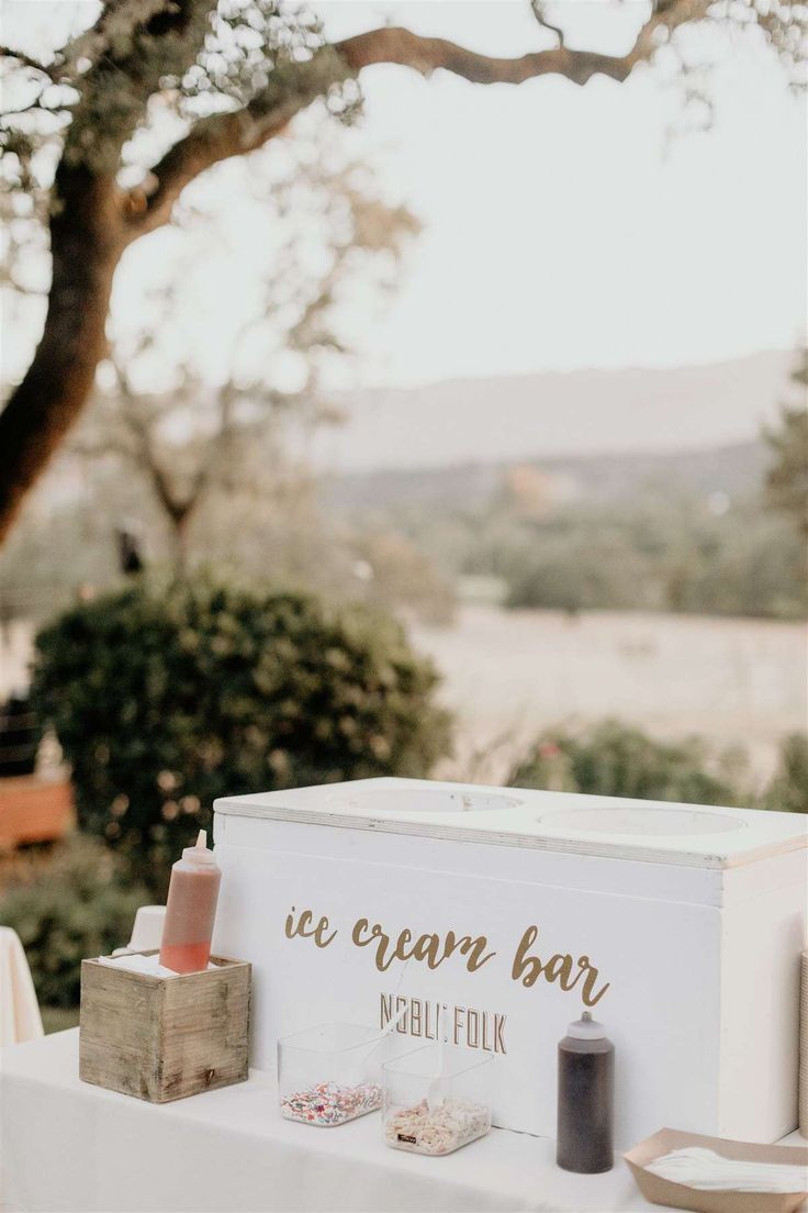 an ice cream bar is set up on a table outside with trees in the background