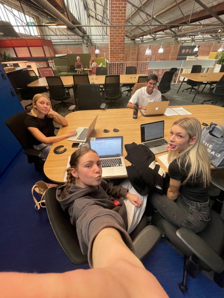 two women sitting at a table with laptops in front of them and one woman pointing to the camera