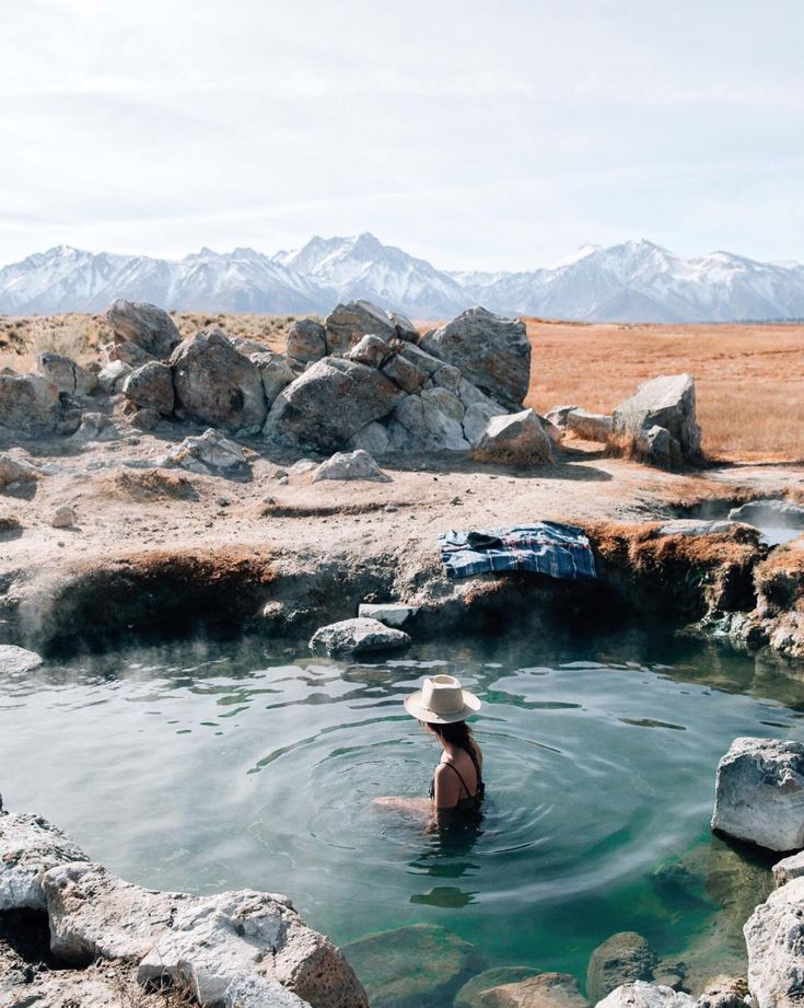 a woman is in the water near some rocks and boulders with mountains in the background