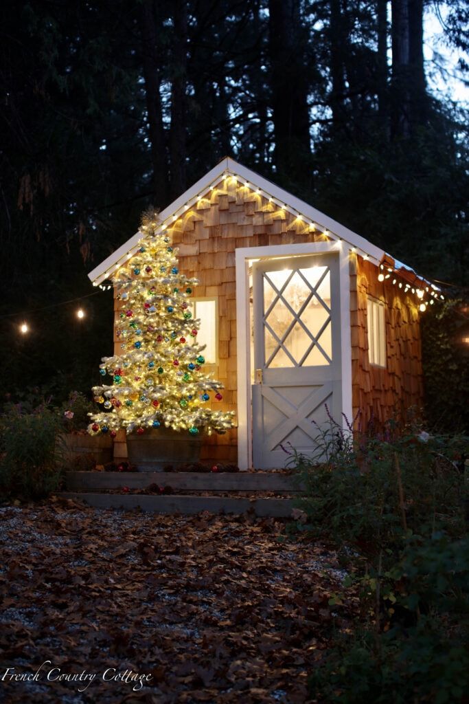 a christmas tree is lit up in front of a small house with lights on it