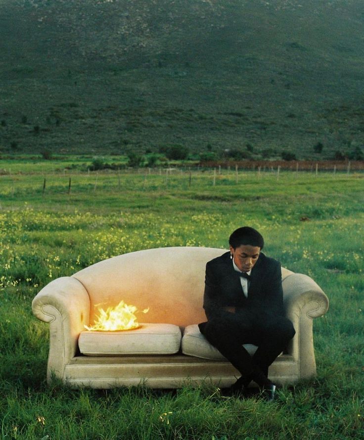 a man sitting on top of a couch in the middle of a field with a fire