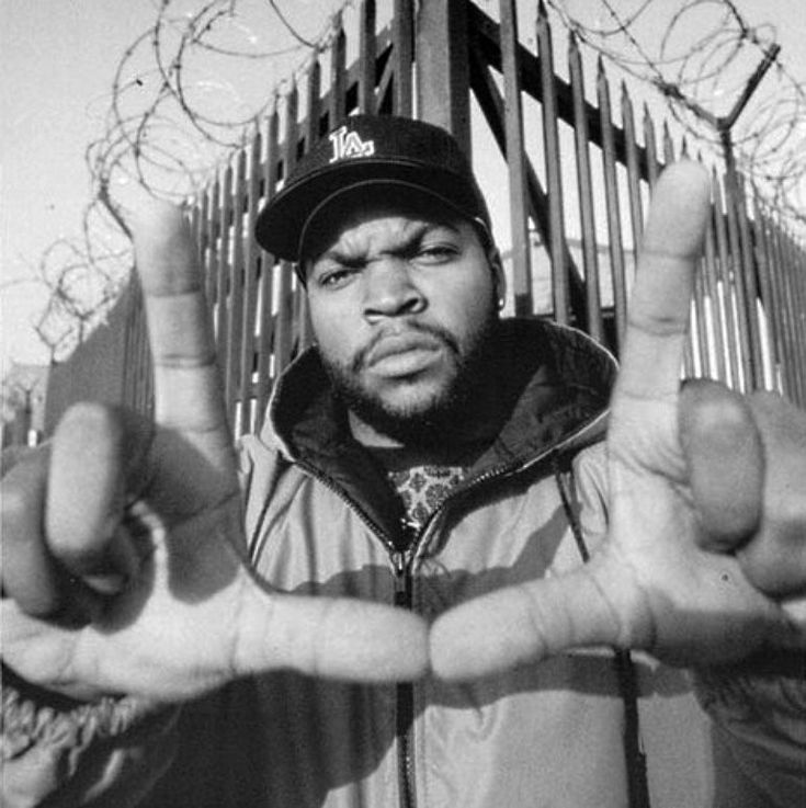 a man standing in front of a fence making the peace sign with his hands while wearing a hat