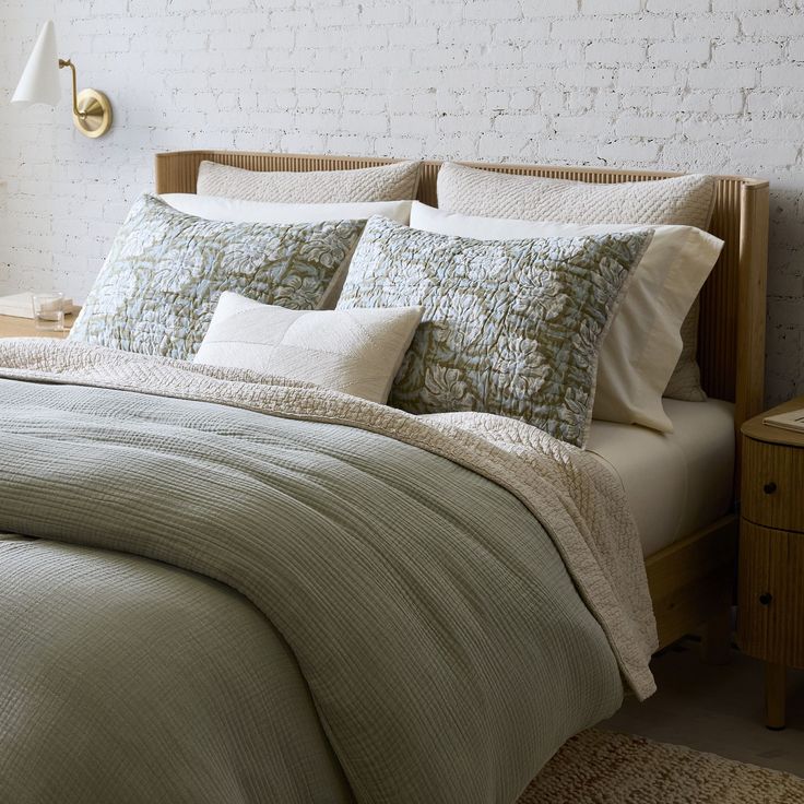 a bed with pillows and blankets on top of it next to a night stand in front of a brick wall