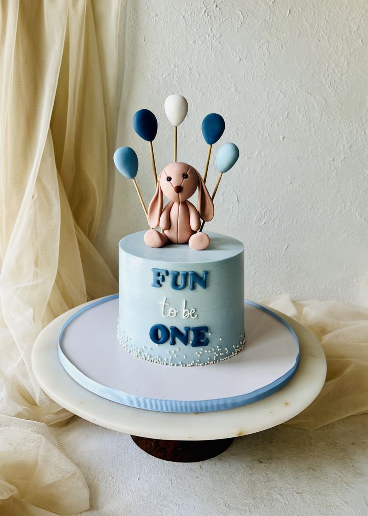 a birthday cake decorated with balloons and a teddy bear on top that says fun to be one