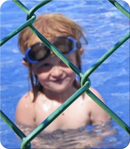 a young child wearing goggles standing in the water behind a chain link fence and smiling