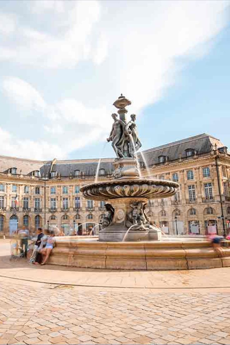 a fountain with people sitting around it in the middle of a courtyard and some buildings