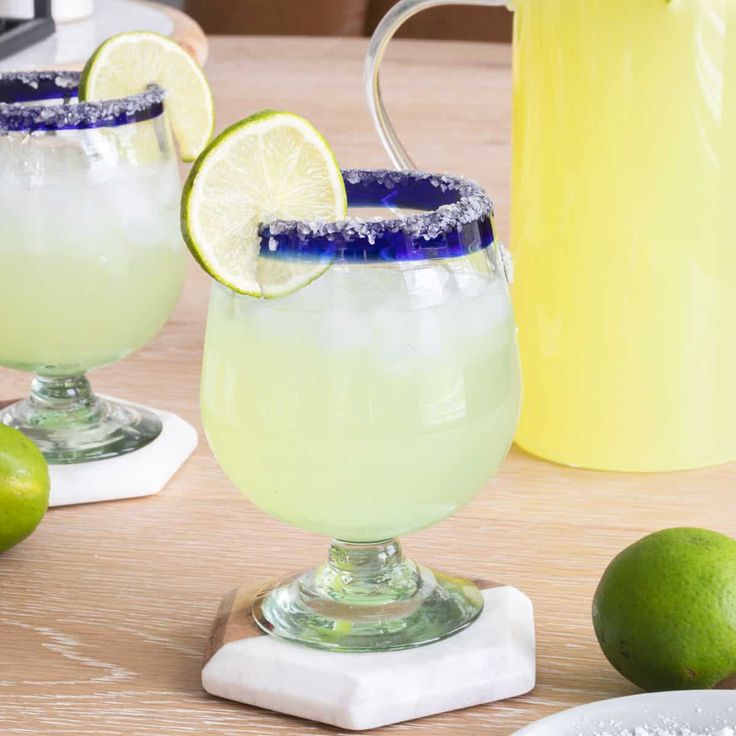 two margaritas sitting on top of a wooden table next to limes and a pitcher