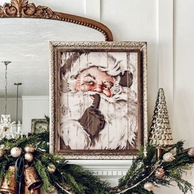 a christmas mantle decorated with greenery, ornaments and an old fashioned framed santa clause