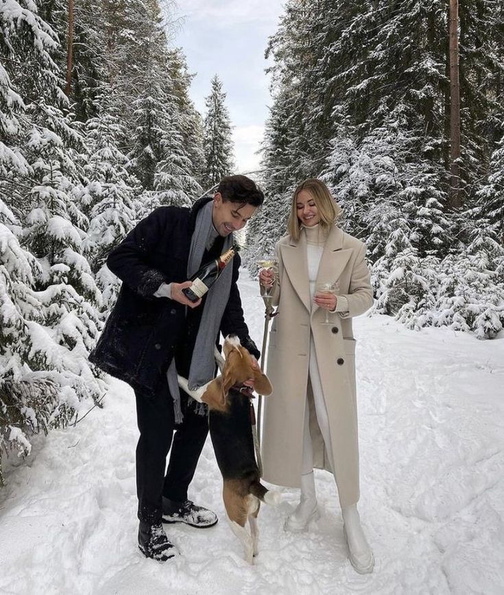 a man and woman are standing in the snow with their dog, holding wine glasses