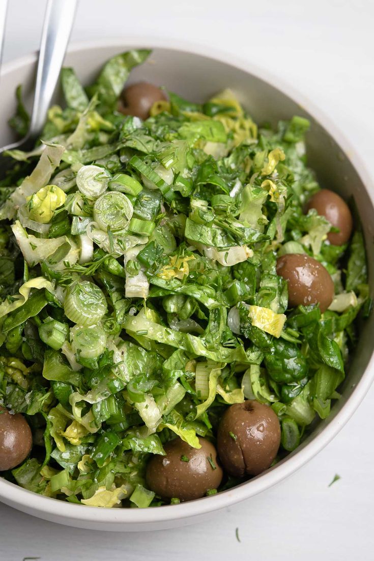 a white bowl filled with lettuce and olives next to a silver spoon