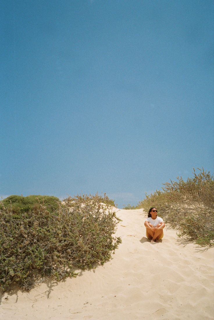 a man sitting on top of a sandy beach under a blue sky with an airplane flying overhead