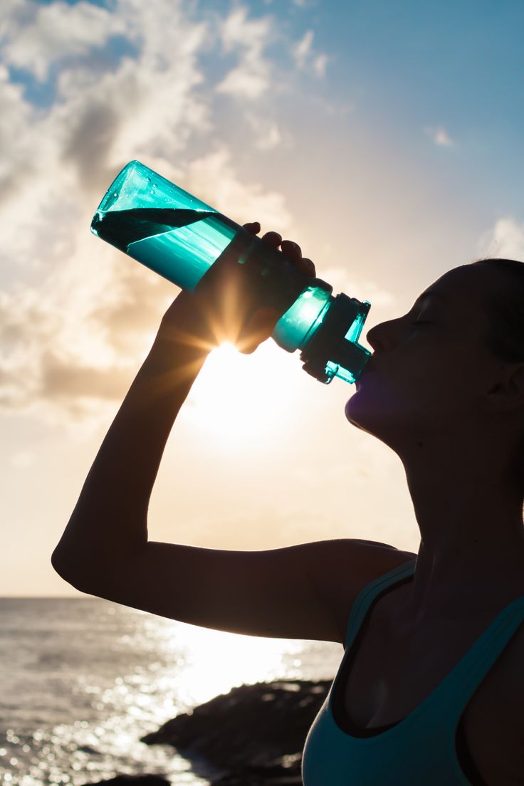 a woman drinking water from a green bottle on the beach at sun set in front of the ocean