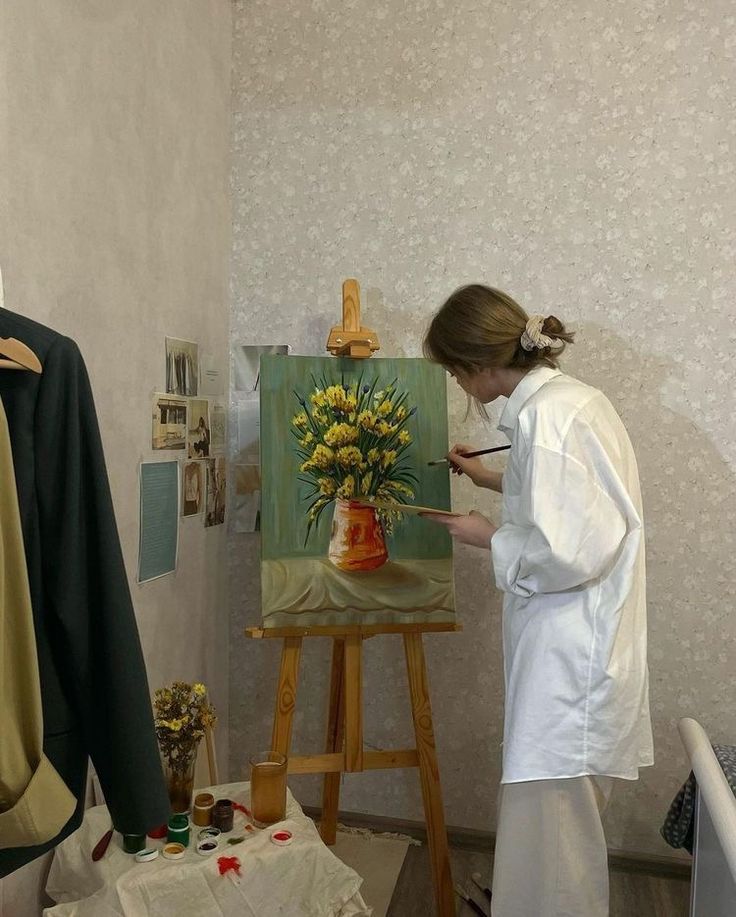 a woman is painting flowers in a vase on an easel next to a coat rack