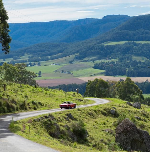 a red car driving down a winding road in the country side with hills and valleys behind it