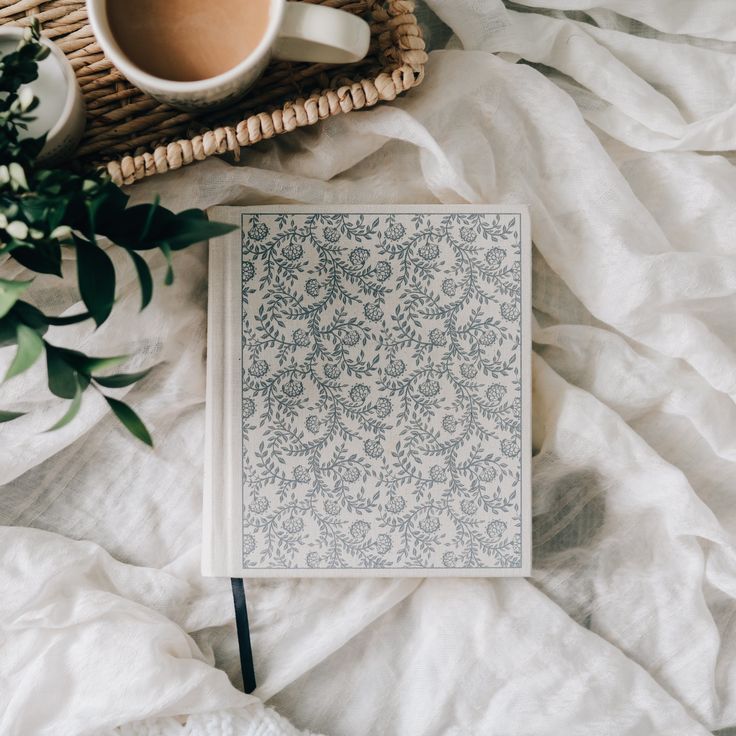 an open notebook sitting on top of a bed next to a cup of coffee and wicker basket