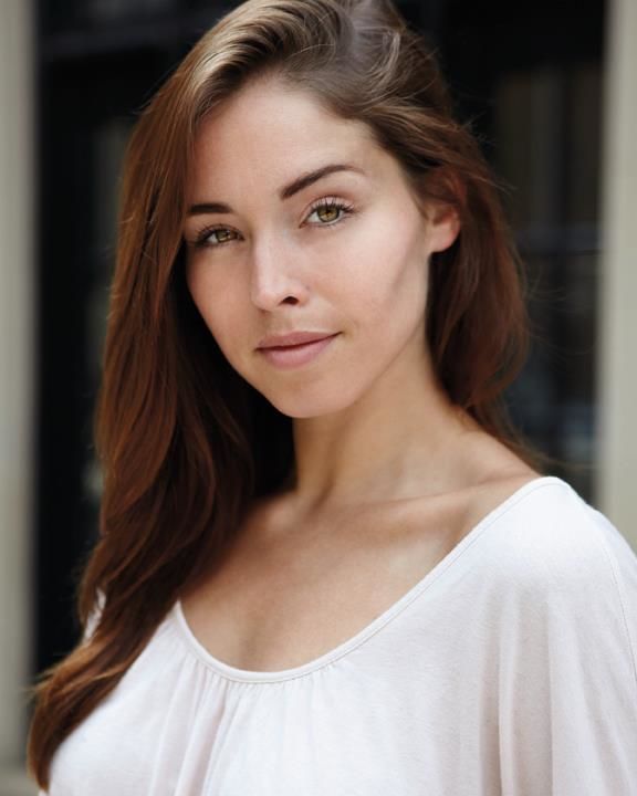 a woman with long brown hair wearing a white shirt and looking at the camera while standing in front of a building