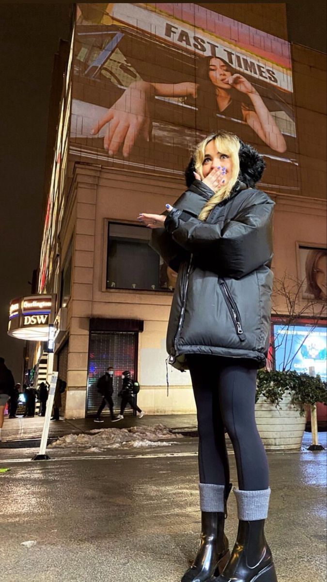 a woman in black jacket and leggings standing on street next to tall building