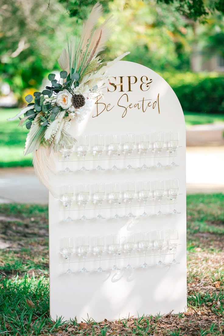 a white sign that says sip and be - seated with flowers on it in the grass