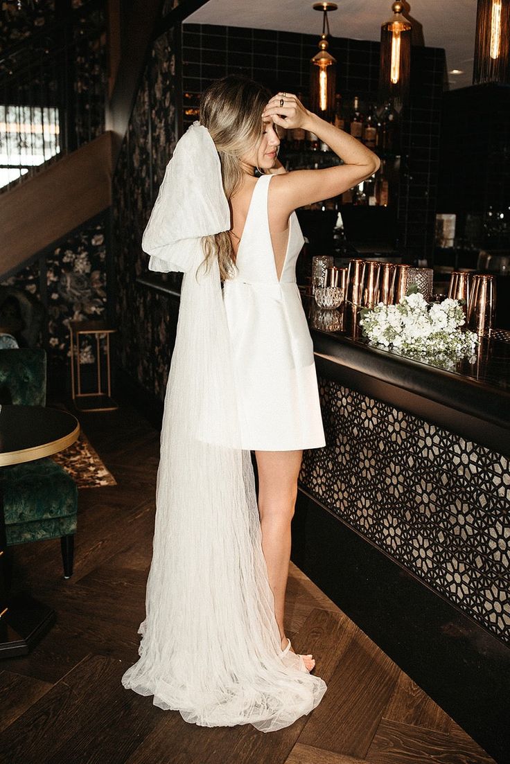 a woman standing in front of a bar wearing a white dress and holding her hair back