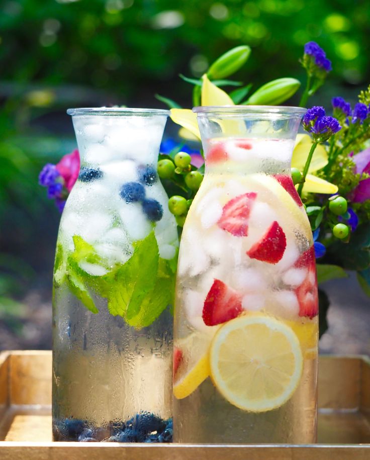 two vases filled with fruit and ice on top of a wooden tray next to flowers