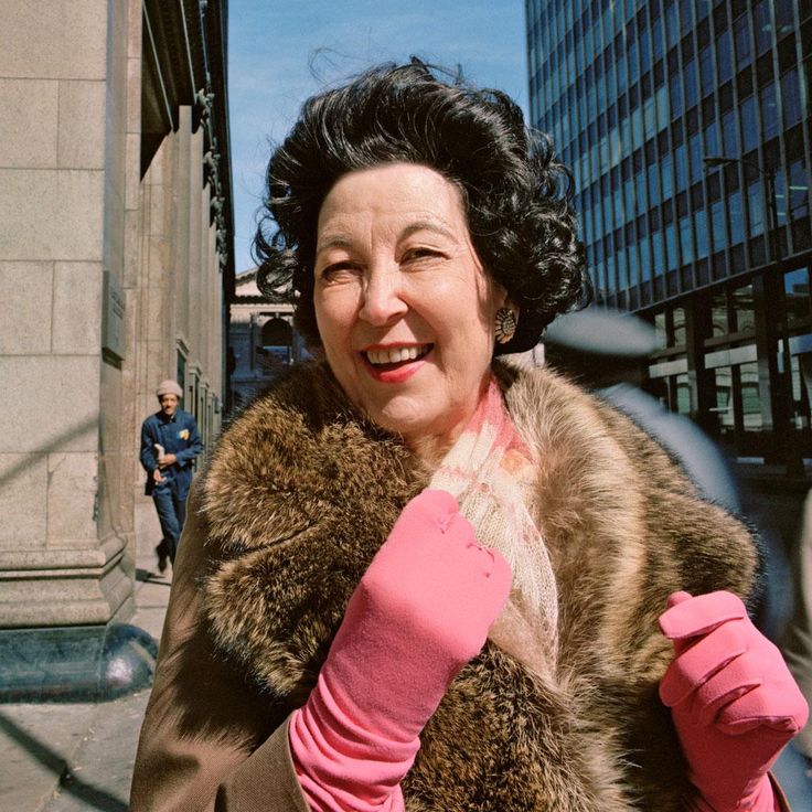 an older woman in pink gloves and fur coat walking down the street with buildings behind her