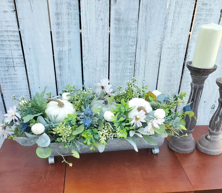 a wooden table topped with a vase filled with white flowers and greenery next to a candle