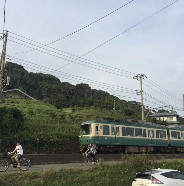 two people riding bikes next to a green and white train on the tracks in front of a hill