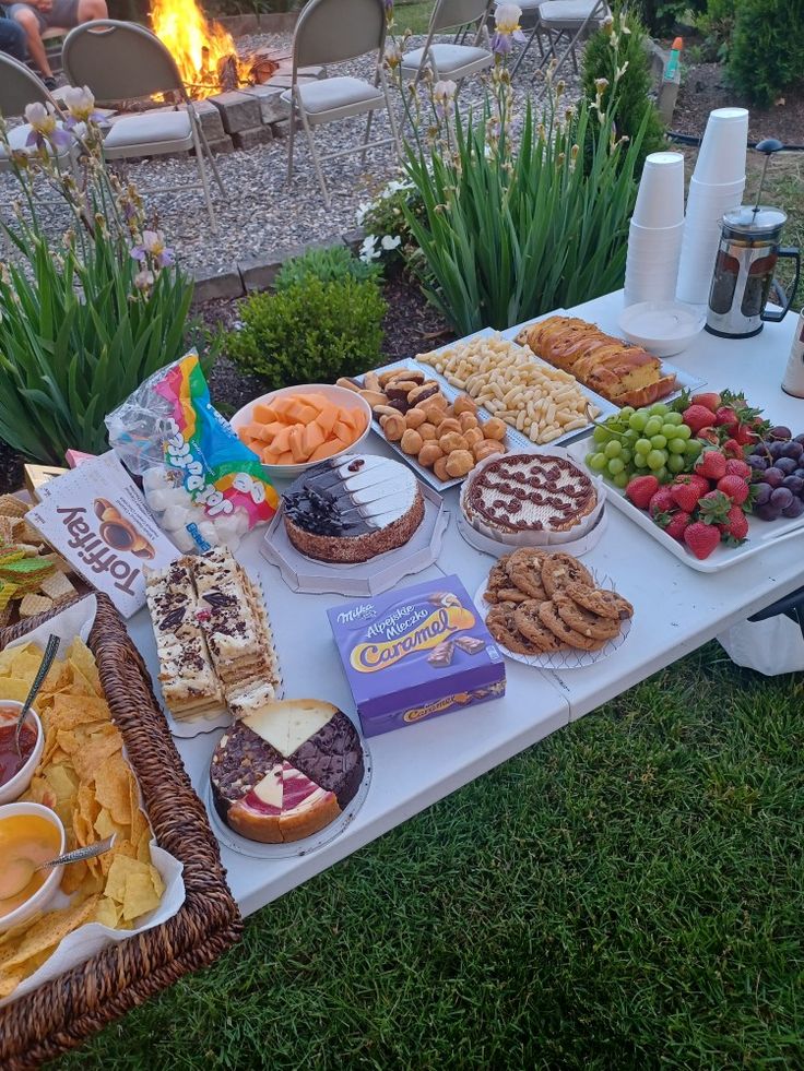 a table filled with food and snacks on top of a grass covered field next to a fire pit