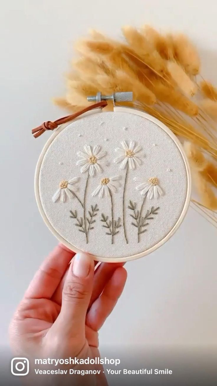 a hand holding up a white embroidery with daisies on it and some brown feathers