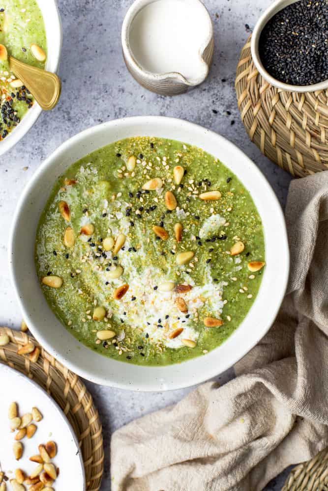 a white bowl filled with green soup next to bowls of seeds and other food items