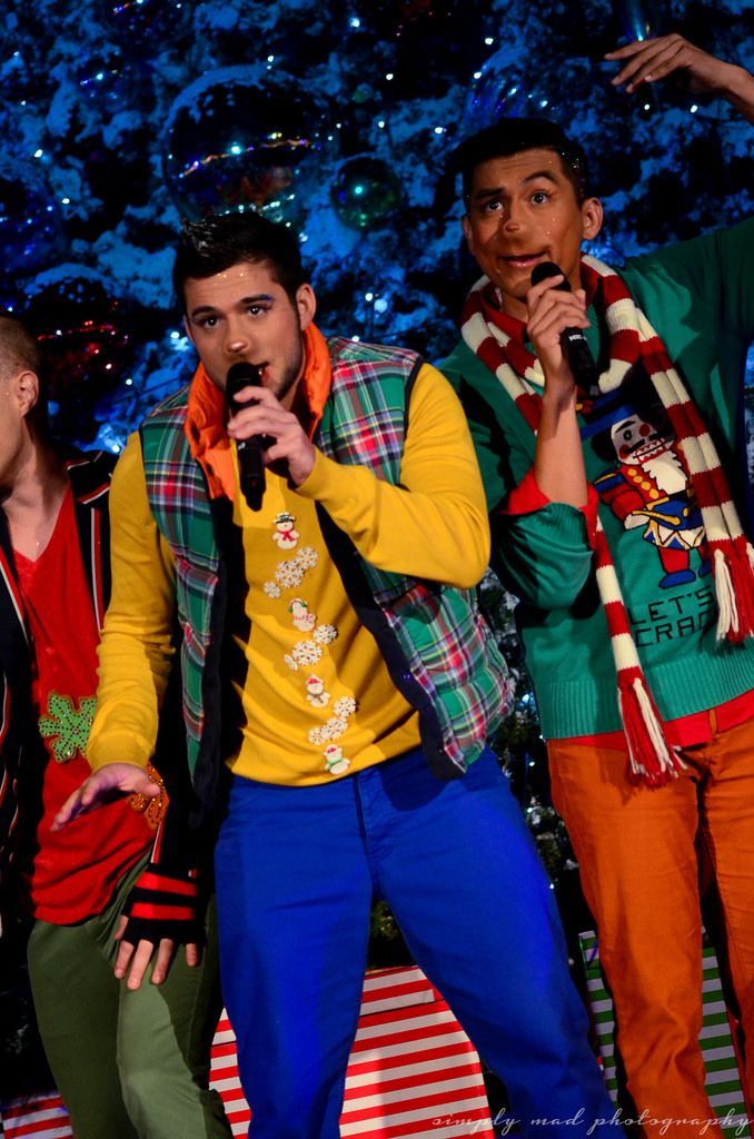 three young men singing into microphones in front of a christmas tree