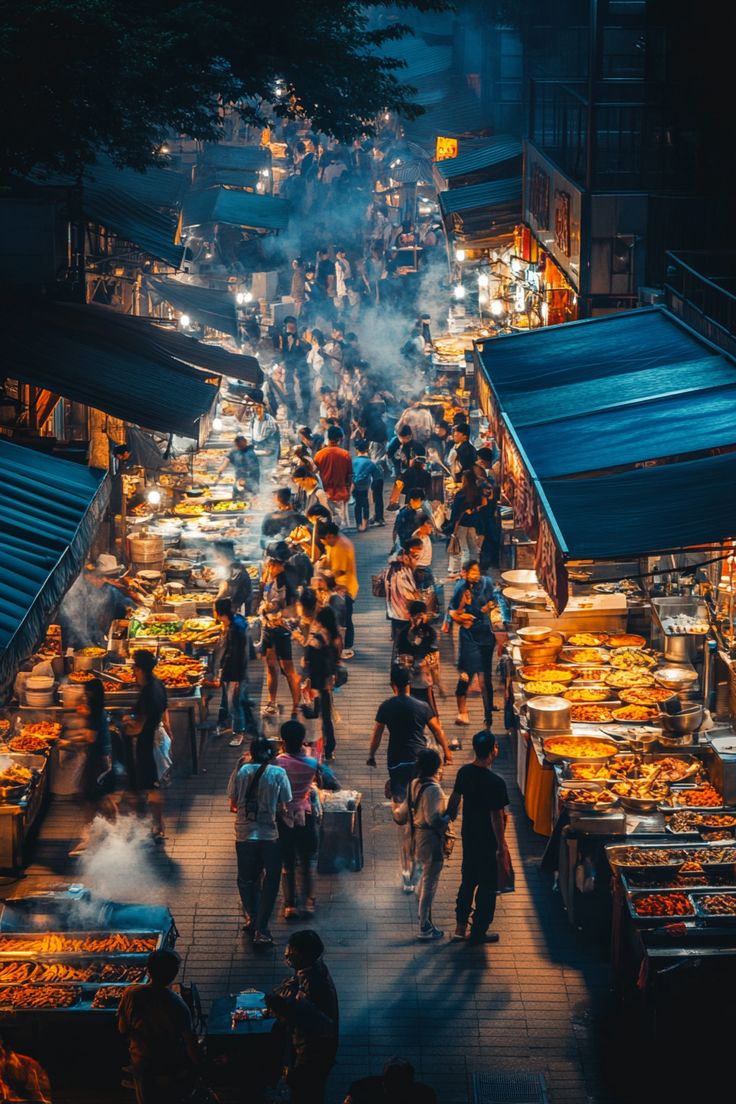 an outdoor market with lots of food and people walking around it at night, in the evening