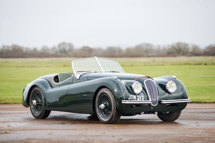 an old green sports car is parked on the pavement in front of a grassy field