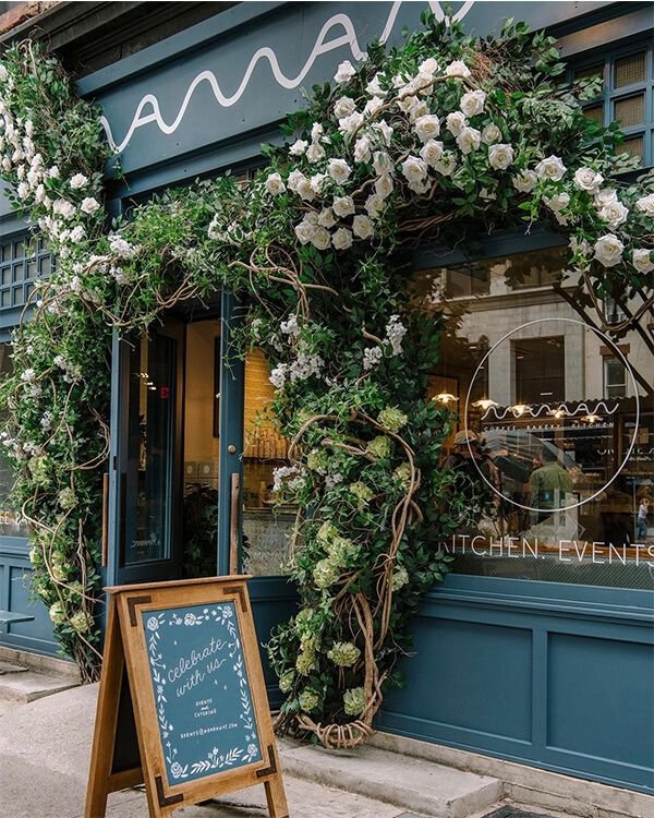 a sign in front of a store with flowers on the outside and greenery around it