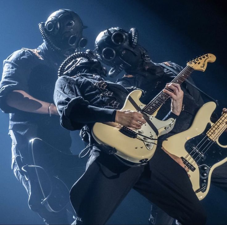 two men in gas masks are playing guitars on stage with one man holding an electric guitar