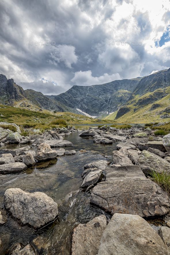 a stream running through a lush green valley surrounded by rocks and grass with mountains in the background